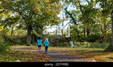 River Tyne, Haddington, East Lothian, Schottland, Großbritannien, 26. Oktober 2020. UK Wetter: Herbst Farben: Sonnenschein mit Herbstblättern beleuchtet in der Sonne, als zwei kleine Mädchen Roller entlang der Uferpromenade fahren Stockfoto