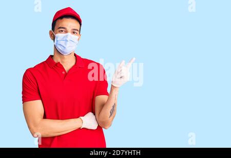 Junge gutaussehende hispanischen Mann trägt Lieferung Uniform und medizinische Maske Lächelnd glücklich zeigt mit Hand und Finger zur Seite Stockfoto