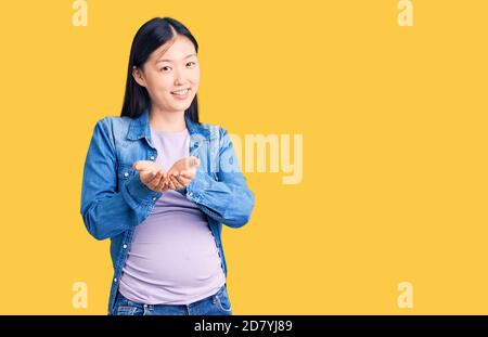 Junge schöne chinesische Frau schwanger erwartet Baby lächelnd mit Händen Handflächen zusammen empfangen oder geben Geste. Halten und Schutz Stockfoto