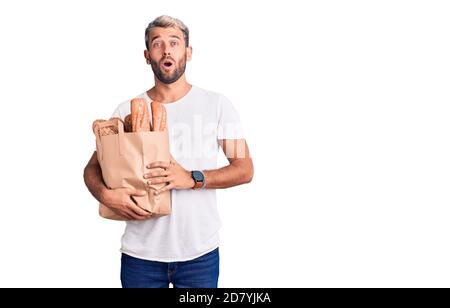 Junge gutaussehende blonde Mann hält Papiertüte mit Brot verängstigt und erstaunt mit offenem Mund für Überraschung, Unglauben Gesicht Stockfoto