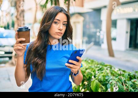 Junge hispanic Frau mit serios Ausdruck mit Smartphone und trinken Take Away Kaffee in der Stadt. Stockfoto