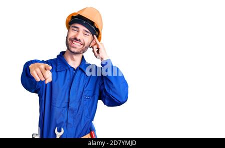 Junger hispanischer Mann, der Arbeiteruniform trägt und lächelt und dabei telefonische Geste macht und auf Sie zeigt. Rufen Sie mich an. Stockfoto