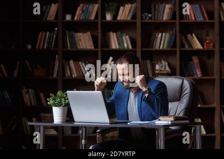 Junger bärtiger Geschäftsmann in einem blauen Anzug freut sich über Erfolg und Sieg beim Blick auf den Laptop-Bildschirm. Mann in der Bürobibliothek Stockfoto