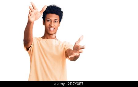 Junger afroamerikanischer Mann in lässiger Kleidung, der die Kamera lächelt und die Arme zur Umarmung offen hat. Fröhlicher Ausdruck, der Glück umarmt. Stockfoto
