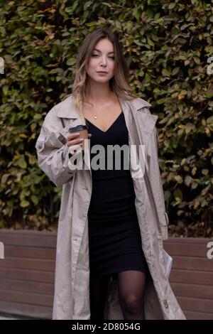 Schöne schlanke junge Brünette in modischen Trenchcoat und schwarzes Kleid mit einer Papiertasse Kaffee. Stockfoto