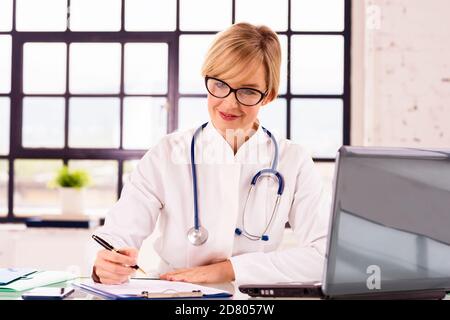 Portrat Aufnahme von lächelnden weiblichen Arzt sitzen am Schreibtisch etwas schreiben, während der Arbeit in Arztpraxis. Stockfoto