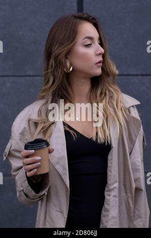 Schöne schlanke junge Brünette in modischen Trenchcoat und schwarzes Kleid mit einer Papiertasse Kaffee. Stockfoto