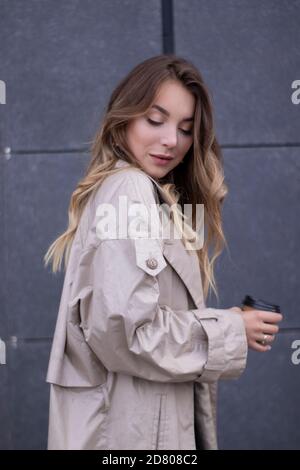 Schöne schlanke junge Brünette in modischen Trenchcoat und schwarzes Kleid mit einer Papiertasse Kaffee. Stockfoto