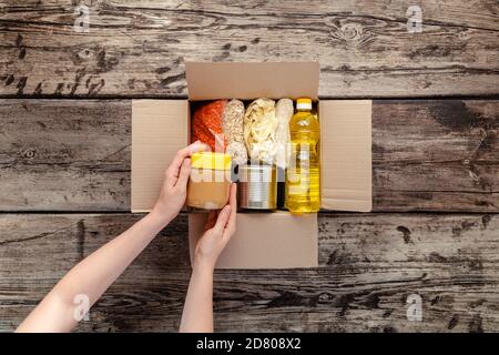 Person Frau, die Spende Nahrungsmittelbox erhält. Weibliche Freiwillige Hände Verpackung Spendenbox mit Lebensmitteln von Grundnahrungsmitteln auf Holztisch. Spenden Sie Essen Stockfoto