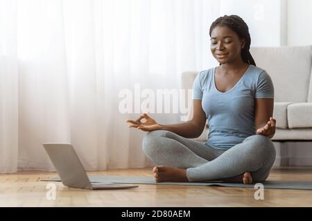 Friedliche afrikanische Dame meditiert zu Hause vor dem Laptop und praktiziert Yoga Stockfoto