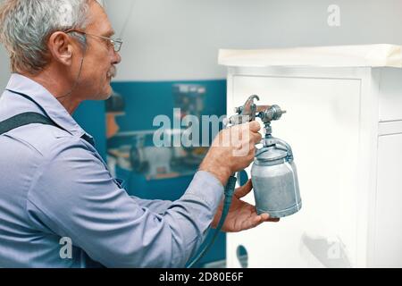 Lackierung von Metallprodukten. Ein älterer Mann malt ein Kabinett mit einem Kompressor Stockfoto