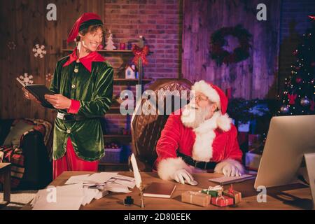 Foto Porträt von santa claus Arbeit auf PC Blick auf Elf hält schwarzes Buch Stockfoto