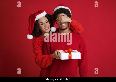 Lächelnd junge afroamerikanische tausendjährige Frau in Weihnachtsmann Hut Schließt die Augen des Mannes in Pullover und gibt ihm Geschenk Stockfoto