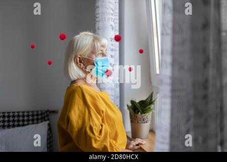 Bleib zu Hause Konzept. Porträt einer erwachsenen 40 50 Jahre alten Frau in einer medizinischen Maske und Hauskleidung steht am Fenster in der Wohnung. Kranke Mädchen und covid 19 Virus fliegt im Zimmer Stockfoto