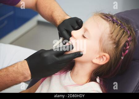 Portrait des kleinen Mädchens hat eine zahnärztliche Untersuchung, während sie im Zahnarztbüro sitzt. Der männliche Zahnarzt überprüft den Mund des jungen Patienten. Stockfoto