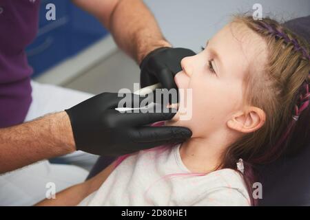 Portrait des kleinen Mädchens hat eine zahnärztliche Untersuchung, während sie im Zahnarztbüro sitzt. Der männliche Zahnarzt überprüft den Mund des jungen Patienten. Stockfoto