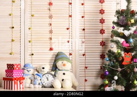 Schneemänner und Teddybären in der Nähe der Uhr und Weihnachtsbaum. Spielzeug auf Kommode auf Holz Wand Hintergrund. Tannenbaum mit Boxen und Dekorationen. Feiern und das Neue Jahr Einrichtung Konzept Stockfoto