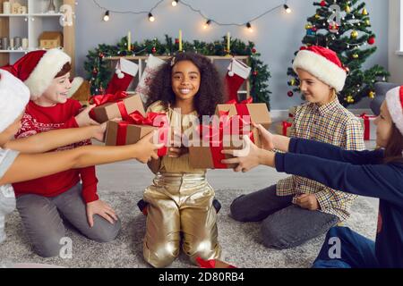 Gruppe von Kindern in Weihnachtsmützen geben Geschenke an ein glückliches afroamerikanisches Mädchen, das Geburtstag hat. Stockfoto