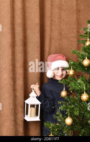 Ein Kind mit einer Laterne in der Hand steht peinlich lächelnd neben einem gekleideten Weihnachtsbaum. Stockfoto