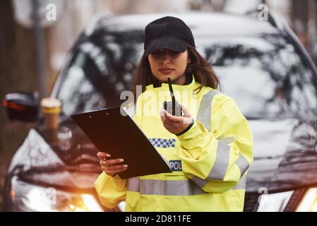 Polizistin in grüner Uniform stehend mit Funksender Und Notizblock gegen Auto Stockfoto