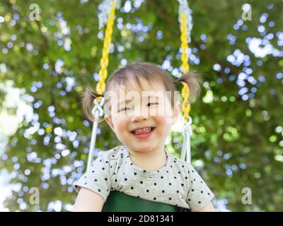 Glücklich lächelnd Kleinkind Mädchen mit Pigtails Schaukeln im Freien unter EIN Baum Stockfoto