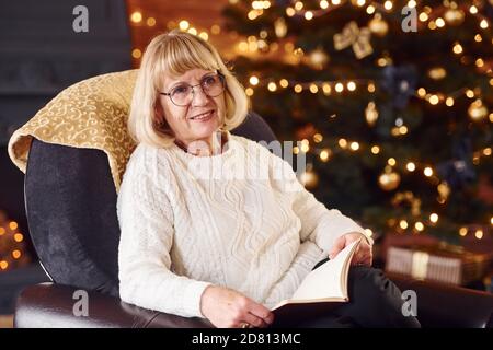 Alte Frau sitzt auf dem Stuhl mit Buch in festlichen Neujahr Zimmer mit weihnachtsdekoration Stockfoto