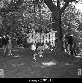 1960er Jahre, historisches Bild von kleinen Kindern stehen als ein kleines Mädchen auf einem Schaukelstuhl sitzen am Geschirr und einem Seil, das an einem Baum gebunden ist, über das Seil über eine Rasenfläche reisen, eine der Aktivitäten in einem Dorf Sommerfete, Fife, Schottland Stockfoto