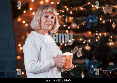 Ältere fröhliche Frau, die mit einer Tasse Getränk in der steht Zimmer mit weihnachtsdekoration Stockfoto