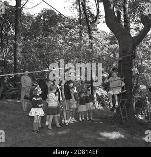 1960er Jahre, historisches Bild von kleinen Kindern stehen, als ein junges Mädchen auf einem Schaukelstuhl sitzen, befestigt an Geschirr und einem Seil, das an einem Baum gebunden ist, über das Seil über eine Rasenfläche zu reisen, eine der Aktivitäten in einem Dorf Sommerfete, Fife, Schottland. Stockfoto