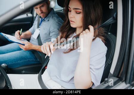 Attraktive junge Frau nervös vor dem Fahrtest, Sicherheitsgurt anlegen und nach unten schauen. Männlicher Selbstausbilder, der weibliche Kursteilnehmer auswertet. Weniger fahren Stockfoto