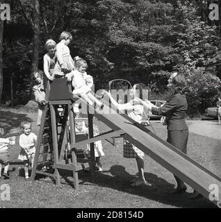 1960er Jahre, draußen auf einem Feld in einem Park, warten kleine Mädchen auf ihre Reihe, um eine hölzerne Rutsche zu gehen, mit ihren Müttern zur Hand, um sie zu fangen, Fife, Schottland. Stockfoto