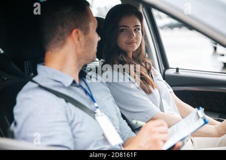 Männlicher Ausbilder lobt attraktive Schülerin für erfolgreiches Fahren. Mann, der etwas in Papier schreibt und mit einer hübschen Frau spricht, die Hi anschaut Stockfoto
