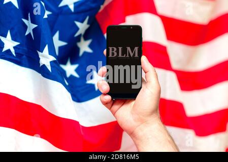 Man's Hand hält ein Handy mit dem Text Abkürzung von Black Lives Matter gegen die verschwommene USA-Flagge. Stockfoto