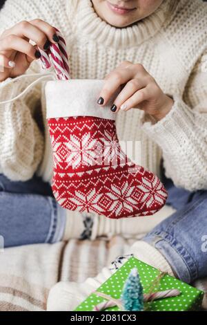 Die junge Frau sitzt auf Karo im kuscheligen Strickpullover aus Wolle und legt gestreiften Lollipop-Stock in die Weihnachtssocke für Geschenke. Vorbereitung auf Winterferien. Aa Stockfoto