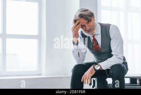 Lächelnder, positiver älterer Mann in Jacke und Krawatte, der drinnen sitzt Im Zimmer gegen das Fenster Stockfoto