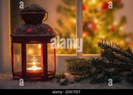 Beleuchtete rote Laterne auf einer Außenwitwowsill bedeckt mit Schnee in einer schneenden Nacht. Ein Weihnachtsbaum im Inneren des Hauses in sichtbar durch das Fenster. Stockfoto