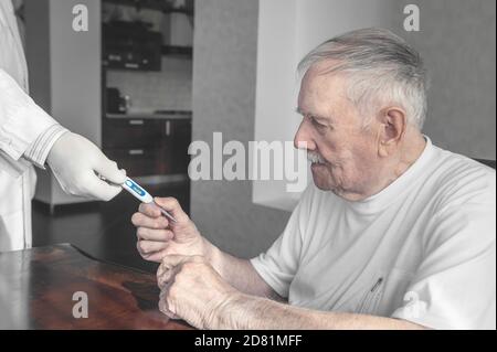 Der Arzt gibt ein Thermometer, um die Temperatur eines älteren kranken Patienten zu messen. Ein grauhaariger alter Mann nimmt dem Arzt das Thermometer aus den Händen Stockfoto