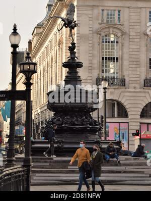 Menschen mit Gesichtsmasken gehen am Shaftesbury Memorial Fountain aka Eros, Piccadilly Circus, London vorbei Stockfoto