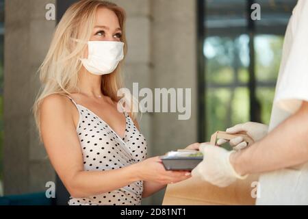 Liebenswert Frau Kunde nehmen ihre Bestellung im Restaurant während Coronavirus, Kellner des Cafés geben Essen in Behälter tragen Schutzhandschuhe, Frau ist in medizinischen Maske Stockfoto
