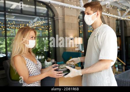 Blonde nette weibliche Klientin machen Ordnung im Restaurant und nehmen Sie es von Kellner in Schutzhandschuhe und Maske. Verschenken Aufträge, Essen in Behältern Stockfoto
