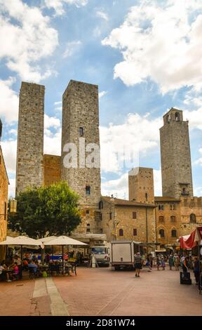 Blick auf die Piazza delle Erbe mit Menschen im Café im Freien und auf dem lokalen Markt unter den berühmten mittelalterlichen Türmen, San Gimignano, Siena, Toskana, Italien Stockfoto