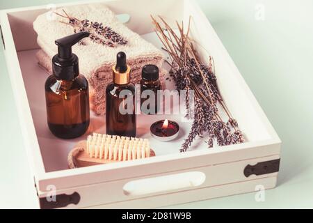 Badkosmetik-Produkte für Körper und Gesicht in braunen Flaschen auf weißem Holz rustikalen Tablett, getrockneten Lavendelblüten, Kerze, Pinsel und Handtücher. Aromatherapie Stockfoto