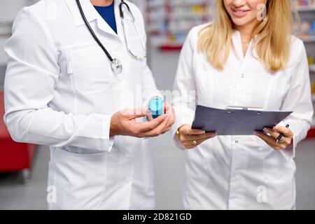 Zwei junge kaukasische Apotheker diskutieren Medikamente, mit einer digitalen Tablette und Dokumente. Mann und Frau in weißem medizinischen Kleid arbeiten in der Apotheke Stockfoto