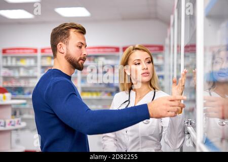 Junge Apothekerin hilft kranken männlichen Kunden Auswahl verschreibungspflichtige Medikamente aus dem Regal, Einkaufen in der Drogerie, unter Beratung von professionellen Chemiker. Konsum, freundliches Personal Konzept Stockfoto