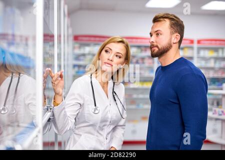 Junge Apothekerin hilft kranken männlichen Kunden Auswahl verschreibungspflichtige Medikamente aus dem Regal, Einkaufen in der Drogerie, unter Beratung von professionellen Chemiker. Konsum, freundliches Personal Konzept Stockfoto