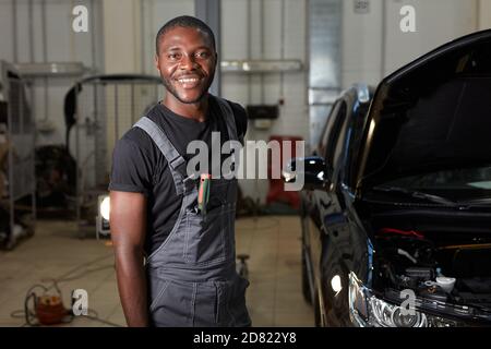 Porträt von lächelnden afrikanischen Auto-Service-Arbeiter am Arbeitsplatz, junge Mann in Uniform Blick auf die Kamera, während der Reparatur eines Autos Stockfoto