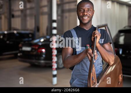 Junge afrikanische Automechaniker bei der Arbeit mit separaten Teil der Maschine, posiert bei der Kamera, gehen zu reparieren Stockfoto