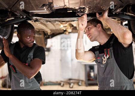 Zwei interracial diverse Mechaniker Überprüfung Auto unten im Auto-Service, kooperieren, arbeiten zusammen Stockfoto