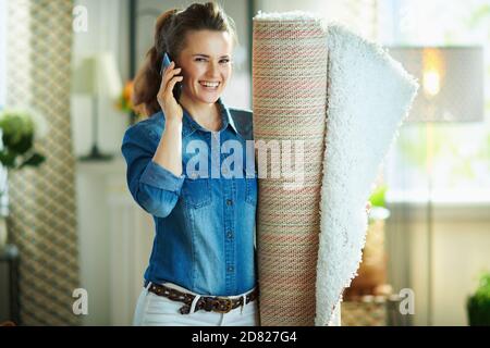 Happy Modern 40 Jahre alte Frau in Jeans-Hemd und weißen Hosen mit weißem Teppich sprechen auf einem Smartphone im modernen Zuhause an sonnigen Tagen. Stockfoto