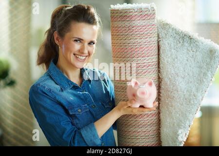 Porträt einer lächelnden 40-jährigen Frau in Jeans-Hemd und weißen Hosen mit weißem Teppich zeigt Sparschwein in der modernen Haus in sonnigen Tag. Stockfoto